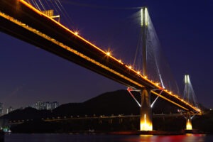 A cable-stayed bridge photographed at night, illuminated by string lights along its cables and deck, with foundation piers rising from dark water and city lights visible in the distance.