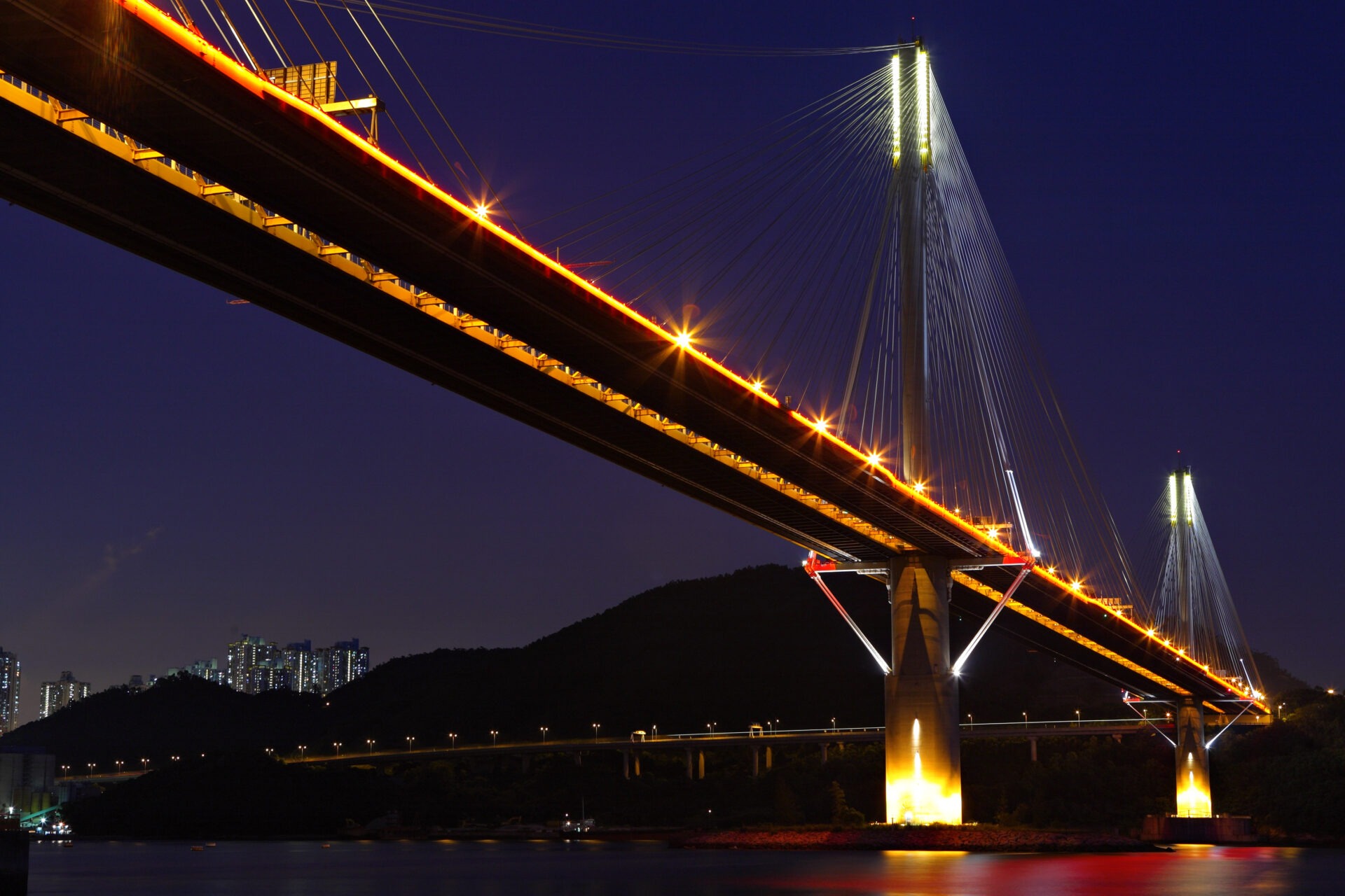 A cable-stayed bridge photographed at night, illuminated by string lights along its cables and deck, with foundation piers rising from dark water and city lights visible in the distance.